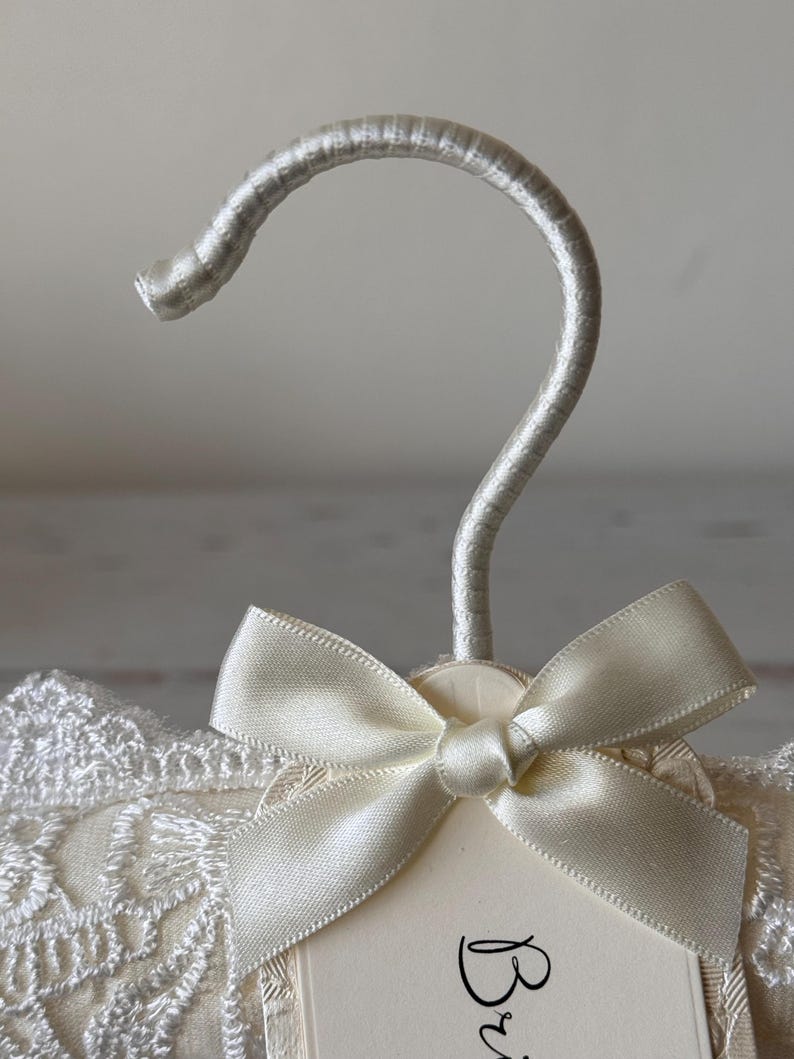 Op de afbeelding: Een gevoerde kledinghanger bekleed met cr&egrave;mekleurige stof met een witte kanten overlay. De hanger heeft een zilverkleurige haak omwikkeld met bijpassende stof en een cr&egrave;mekleurige satijnen strik aan de basis van de haak, met een label Bride.