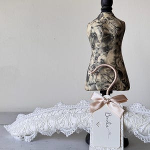 Op de afbeelding: Een witte gewatteerde kledinghanger bedekt met ingewikkeld kant, voorzien van een ros&eacute;gouden haak en een rechthoekig label met het woord Bride in zwarte letters. Het label is versierd met een kleine satijnen strik en een hartvormige uitsparing.