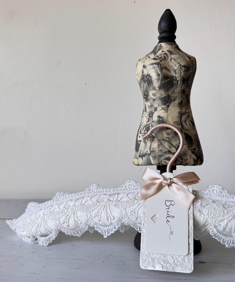 Op de afbeelding: Een witte gewatteerde kledinghanger bedekt met ingewikkeld kant, voorzien van een ros&eacute;gouden haak en een rechthoekig label met het woord Bride in zwarte letters. Het label is versierd met een kleine satijnen strik en een hartvormige uitsparing.