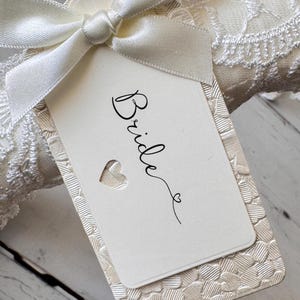 Op de afbeelding: Een rechthoekig gebroken wit label met het woord Bride in zwarte cursieve letters en een klein hartje. Het label ligt op een gestructureerd cr&egrave;mekleurig karton met een hartjespatroon en is vastgemaakt met een ivoorkleurig satijnen lint.