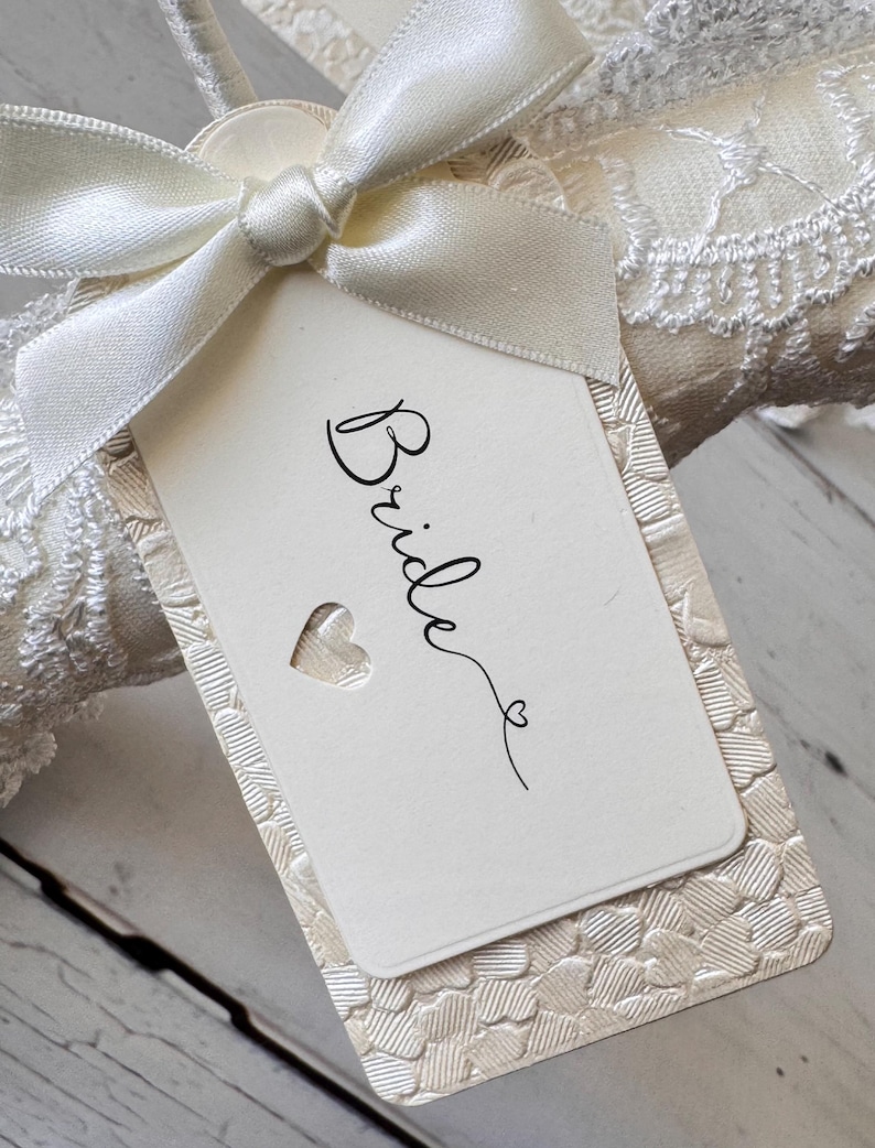 Op de afbeelding: Een rechthoekig gebroken wit label met het woord Bride in zwarte cursieve letters en een klein hartje. Het label ligt op een gestructureerd cr&egrave;mekleurig karton met een hartjespatroon en is vastgemaakt met een ivoorkleurig satijnen lint.