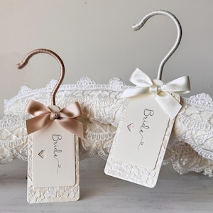 Op de afbeelding: Twee rechthoekige labels met afgeronde hoeken, elk met een kleine hartvormige uitsparing en het woord Bride in zwarte schrijfletters. E&eacute;n label heeft een ros&eacute;gouden haak en lint, de andere een zilveren haak en wit lint, met kanttextuur.