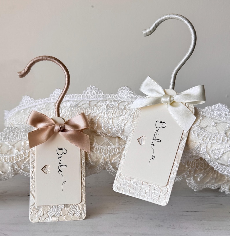 Op de afbeelding: Twee rechthoekige labels met afgeronde hoeken, elk met een kleine hartvormige uitsparing en het woord Bride in zwarte schrijfletters. E&eacute;n label heeft een ros&eacute;gouden haak en lint, de andere een zilveren haak en wit lint, met kanttextuur.