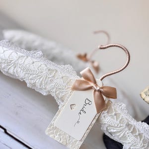 Op de afbeelding: Een witte, gevoerde kledinghanger omwikkeld met gedetailleerd wit kant. De hanger heeft een ros&eacute;gouden metalen haak en een rechthoekig gebroken wit label vastgemaakt met een klein satijnen lint. Op het label staat het woord Bride en een hartje.