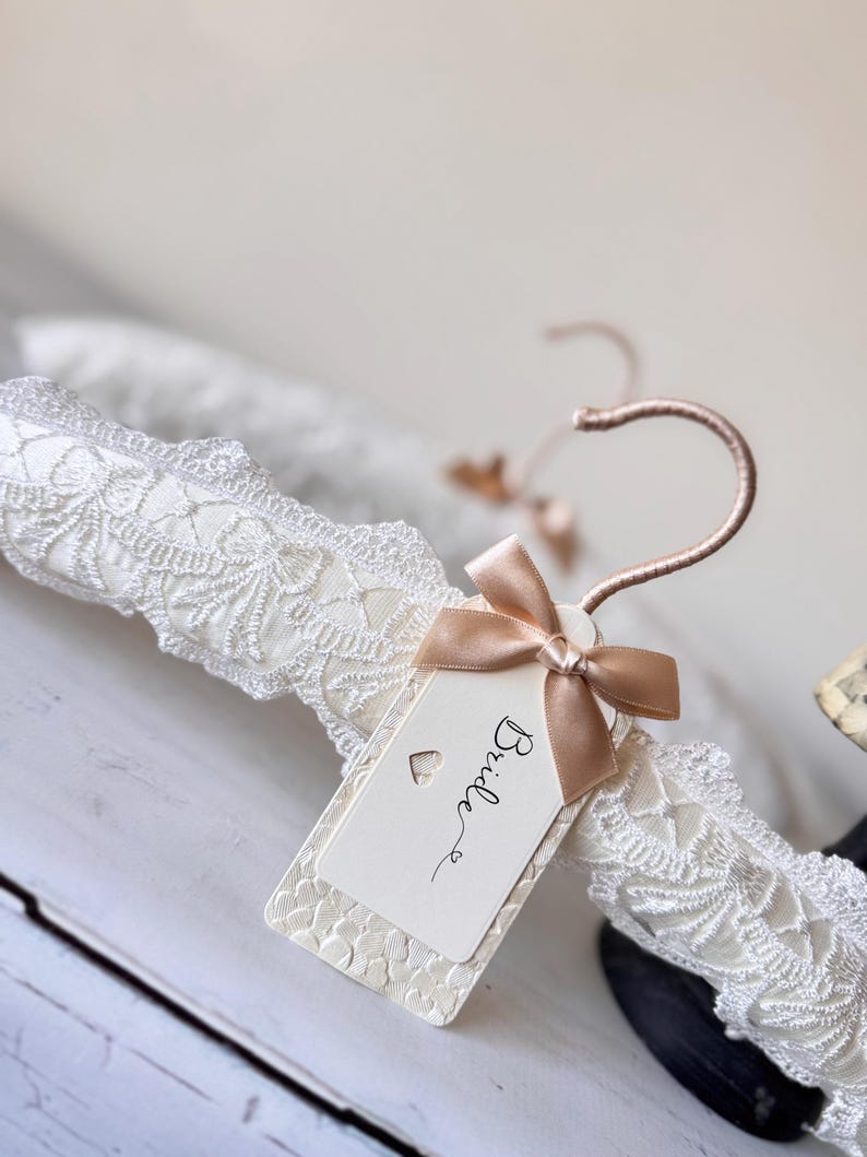 Op de afbeelding: Een witte, gevoerde kledinghanger omwikkeld met gedetailleerd wit kant. De hanger heeft een ros&eacute;gouden metalen haak en een rechthoekig gebroken wit label vastgemaakt met een klein satijnen lint. Op het label staat het woord Bride en een hartje.