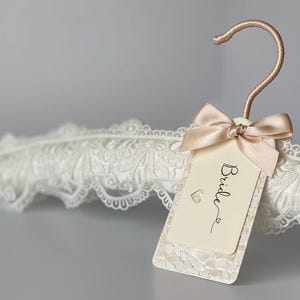 Op de afbeelding: Een met wit kant beklede kledinghanger met een ros&eacute;gouden haak. Een cr&egrave;mekleurig rechthoekig label hangt aan de haak met een lichtbeige satijnen strik. Op het label staat het woord Bride in zwarte schrijfletters en een klein hartje afgebeeld.