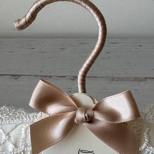 Op de afbeelding: Een gevoerde kledinghanger met een champagnekleurige satijnen haak en een bijpassende satijnen strik in het midden. De hanger is omwikkeld met cr&egrave;mekleurige stof met witte kanten rand en bevat een label met de gedeeltelijke tekst Bride.
