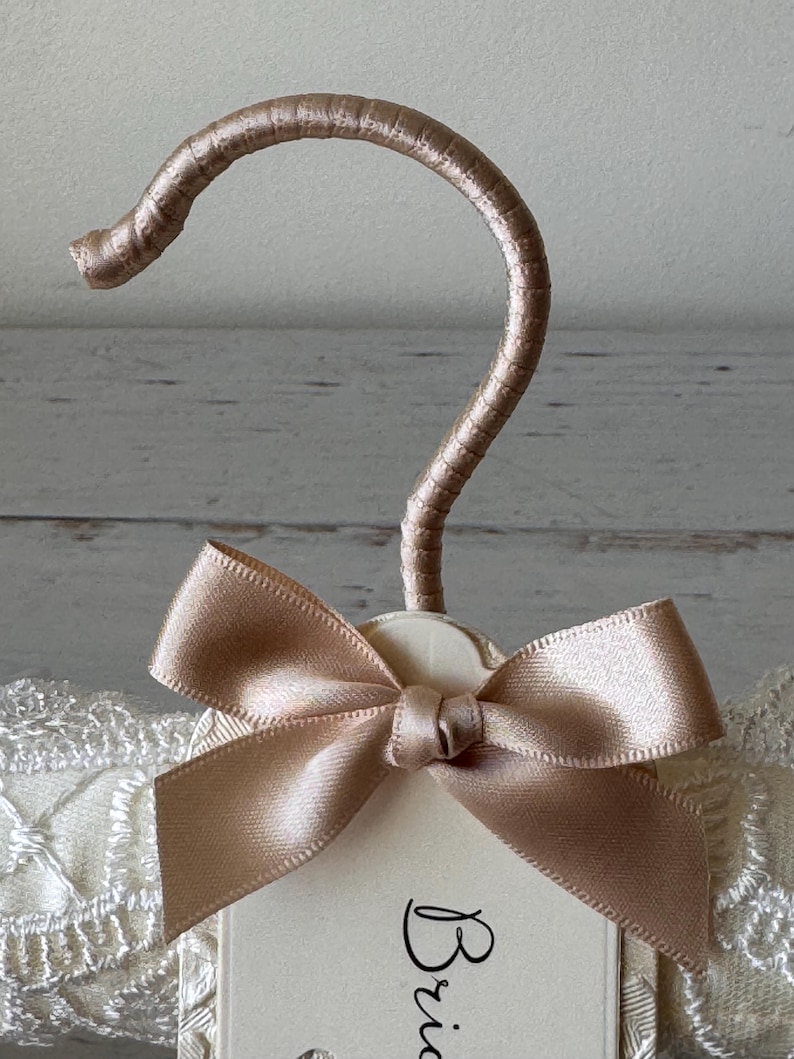 Op de afbeelding: Een gevoerde kledinghanger met een champagnekleurige satijnen haak en een bijpassende satijnen strik in het midden. De hanger is omwikkeld met cr&egrave;mekleurige stof met witte kanten rand en bevat een label met de gedeeltelijke tekst Bride.