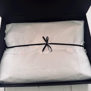 Op de afbeelding: Een rechthoekige zwarte geschenkdoos met een in wit vloeipapier verpakt item. Het papier is vastgezet met een dun zwart lint dat in het midden tot een strik is gebonden, met een kleine ronde sticker met tekst onder de knoop van het lint.