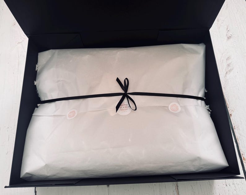 Op de afbeelding: Een rechthoekige zwarte geschenkdoos met een in wit vloeipapier verpakt item. Het papier is vastgezet met een dun zwart lint dat in het midden tot een strik is gebonden, met een kleine ronde sticker met tekst onder de knoop van het lint.