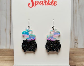 May include: A pair of dangle earrings featuring a black glitter cauldron design with iridescent smoke. The earrings are displayed on a white stand with the text "Sugar and Sparkle" in blue and red.
