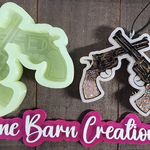 May include: The image showcases a light green silicone mold of two crossed revolvers, alongside a finished pendant with a black cord. The pendant features two crossed revolvers with glitter handles and a black and gold finish. The text "Pine Barn Creations" is also visible.