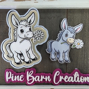May include: Two donkey-shaped stickers, one white and one gray, each holding a daisy. The white donkey is outlined in tan and black. The stickers are above a magenta sign that reads "Pine Barn Creations" in white script.