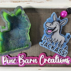 May include: Two donkey-shaped air fresheners. One is a mold with the text "DON'T BE A JACKASS!" and the other is a finished product with the same text. The air freshener has a pink bead and black beads on a string.