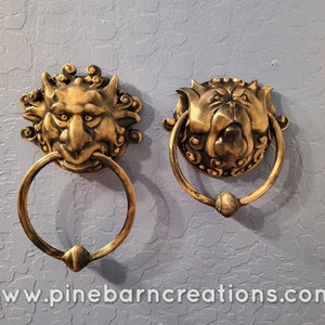 May include: Two antique brass door knockers. One features a devilish face with horns, the other a bulldog-like face. Both have a ring for knocking. The knockers are mounted on a textured, light blue wall.