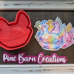 May include: A red plastic chicken-shaped cookie cutter next to a sticker sheet of colorful chickens in nests. The sticker sheet features chickens in various colors, including blue, yellow, and pink. The text "Pine Barn Creations" is also visible.