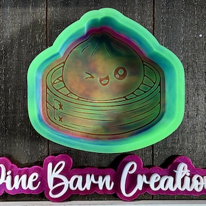 May include: A green and pink dumpling-shaped cookie cutter with a winking face design. The cutter is surrounded by a green border and features a detailed dumpling design. The text "Pine Barn Creations" is below the cutter.