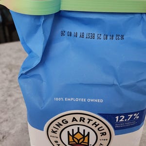 May include: A blue bag of King Arthur Unbleached Bread Flour with a colorful clip. The bag has the text "100% Employee Owned" and "12.7% Gluten-Forming Protein Content". The clip is pastel-colored.