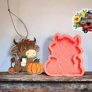 May include: A brown and white cartoon bull with a pumpkin and a cup of coffee, holding a pumpkin. The bull is a Christmas ornament. The ornament is next to a pink silicone mold of the bull.