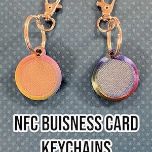 May include: Two NFC business card keychains with gold-colored metal rings and clips. The keychains feature round, iridescent discs with a textured surface. The text "NFC BUSINESS CARD KEYCHAINS" is printed in black on a white background.