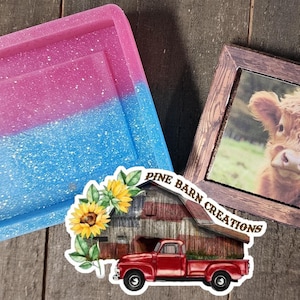 May include: A pink and blue glitter resin tray with a double border. The tray is next to a wooden picture frame with a photo of a brown and white Highland cow. The picture frame has a brown wooden hanger and the text "Pine Barn Creations" on the bottom right corner.