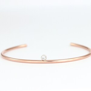 May include: A rose gold cuff bracelet with a small, round, clear gemstone set in the center. The bracelet has a simple, minimalist design and is set against a white background.