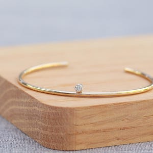 May include: A gold-tone open cuff bracelet with a small, round, clear gemstone in the center. The bracelet is resting on a light brown wooden surface. The jewelry is simple and elegant.