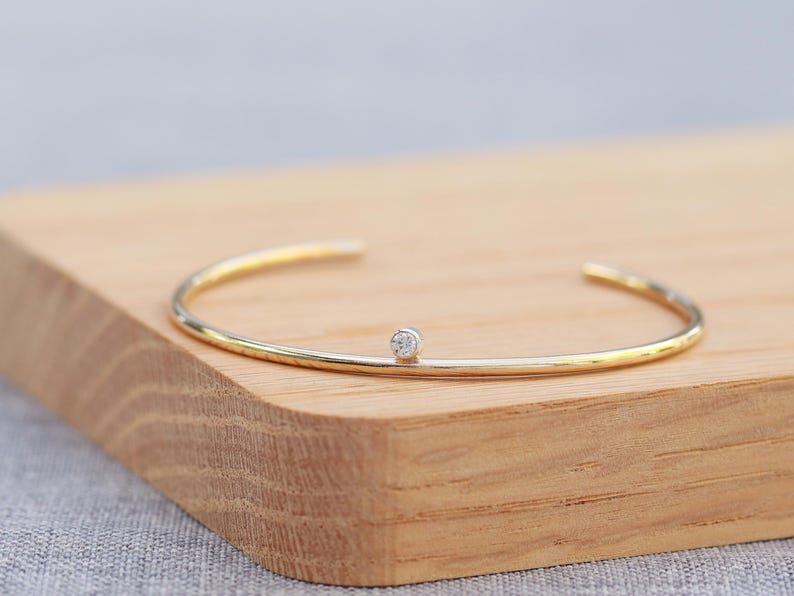 May include: A gold-tone open cuff bracelet with a small, round, clear gemstone in the center. The bracelet is resting on a light brown wooden surface. The jewelry is simple and elegant.