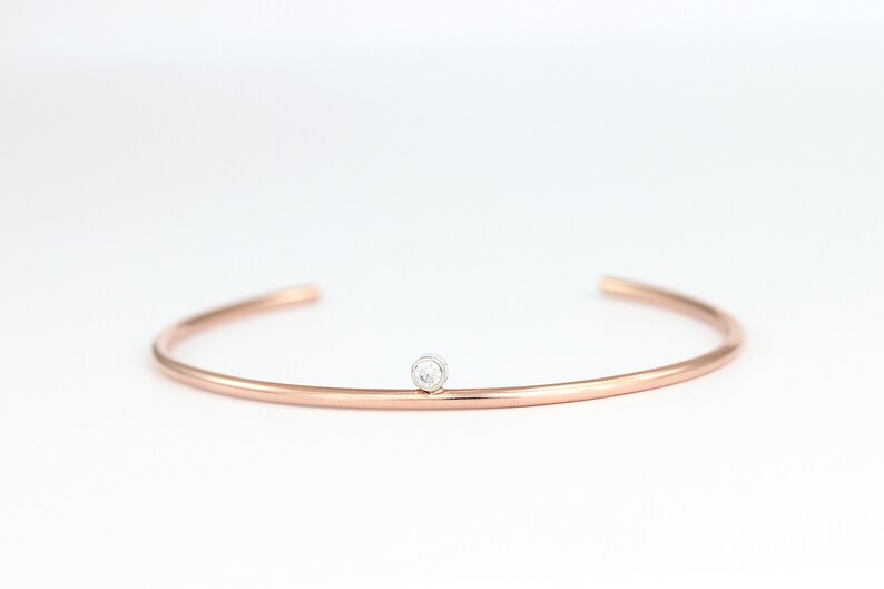 May include: A rose gold cuff bracelet with a small, round, clear gemstone set in the center. The bracelet has a simple, minimalist design and is set against a white background.