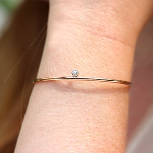May include: A delicate gold-tone bracelet with a small, round, clear gemstone. The bracelet is a thin, minimalist design, and is worn on a wrist. The gemstone is set in a simple, unobtrusive setting.