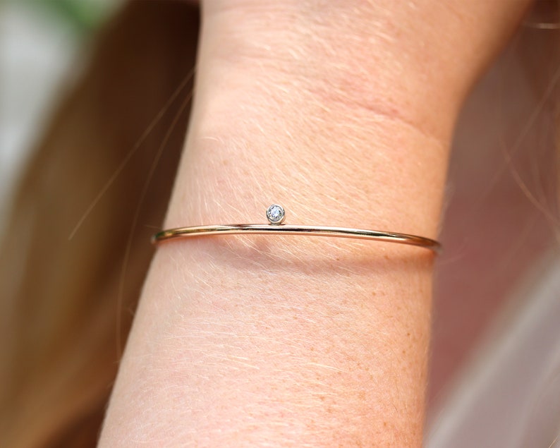 May include: A delicate gold-tone bracelet with a small, round, clear gemstone. The bracelet is a thin, minimalist design, and is worn on a wrist. The gemstone is set in a simple, unobtrusive setting.