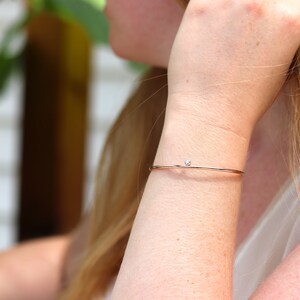 May include: A delicate rose gold bangle bracelet with a small, round, clear gemstone. The bracelet is thin and minimalist, designed to be worn on the wrist. The gemstone is centered on the bracelet.
