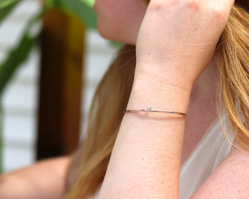 May include: A delicate rose gold bangle bracelet with a small, round, clear gemstone. The bracelet is thin and minimalist, designed to be worn on the wrist. The gemstone is centered on the bracelet.
