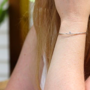 May include: A delicate silver bracelet with a small, clear gemstone. The thin, minimalist design of the bracelet is shown on a person's wrist, highlighting its simple elegance. The gemstone is centered on the bracelet.
