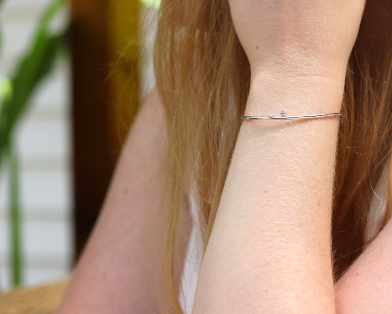 May include: A delicate silver bracelet with a small, clear gemstone. The thin, minimalist design of the bracelet is shown on a person's wrist, highlighting its simple elegance. The gemstone is centered on the bracelet.