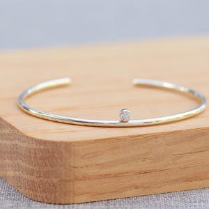 May include: A silver and gold-tone cuff bracelet with a small, round, clear gemstone. The bracelet is open at the ends and rests on a wooden surface. The jewelry is simple and elegant.