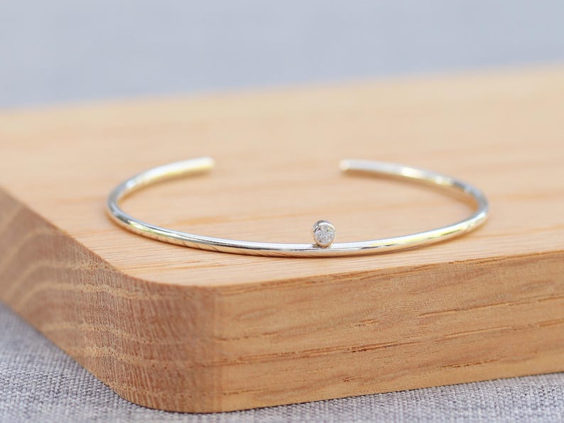 May include: A silver and gold-tone cuff bracelet with a small, round, clear gemstone. The bracelet is open at the ends and rests on a wooden surface. The jewelry is simple and elegant.