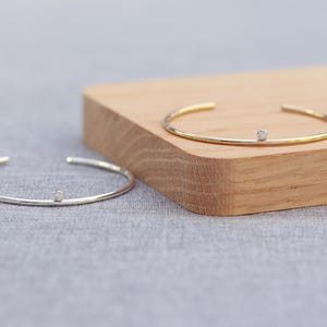 May include: Two open cuff bracelets. One is silver-toned, the other gold-toned. Each bracelet features a small, round, clear gemstone. The bracelets are displayed on a wooden block and a gray surface.