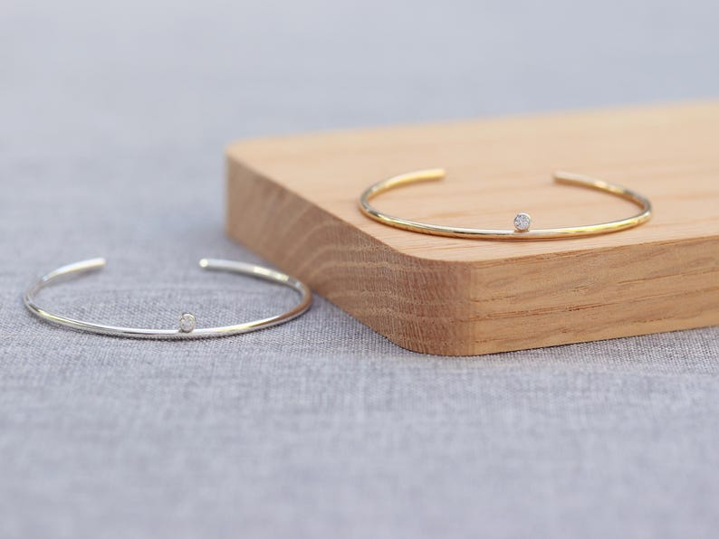 May include: Two open cuff bracelets. One is silver-toned, the other gold-toned. Each bracelet features a small, round, clear gemstone. The bracelets are displayed on a wooden block and a gray surface.