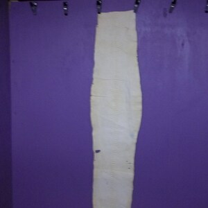May include: A long, off-white piece of fabric hangs vertically against a purple wall. The fabric is textured and appears to be made of a natural material. Metal clips hold the fabric in place at the top. The wall is a solid, vibrant purple.