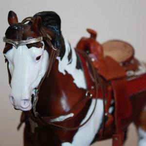 Puede incluir: Un modelo de caballo pintado de marr&oacute;n y blanco con una crin y cola negras. El caballo lleva una brida de cuero marr&oacute;n con una cinta de cabeza plateada y una silla de montar de cuero marr&oacute;n con detalles rojos y marrones.