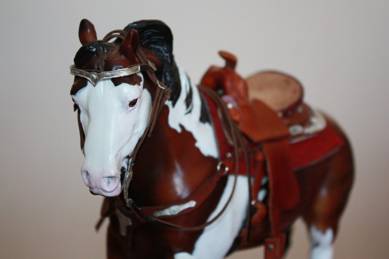 Puede incluir: Un modelo de caballo pintado de marr&oacute;n y blanco con una crin y cola negras. El caballo lleva una brida de cuero marr&oacute;n con una cinta de cabeza plateada y una silla de montar de cuero marr&oacute;n con detalles rojos y marrones.