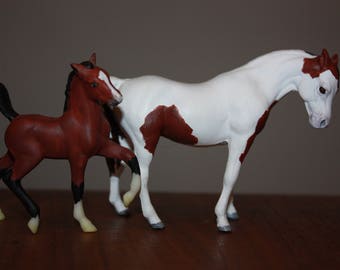 Puede incluir: Una figurita de caballo pintada de marrón y blanco de pie junto a una figurita de caballo más pequeña pintada de marrón y negro. Ambas figuritas están hechas de plástico y tienen detalles realistas.