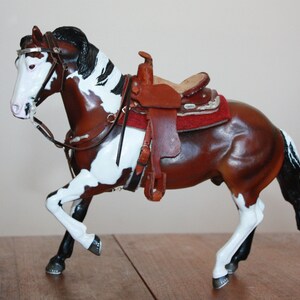 Puede incluir: Una figurita de caballo pintada de marr&oacute;n y blanco con una crin y cola negras. El caballo lleva una silla de montar de cuero marr&oacute;n con una almohadilla roja y detalles plateados.
