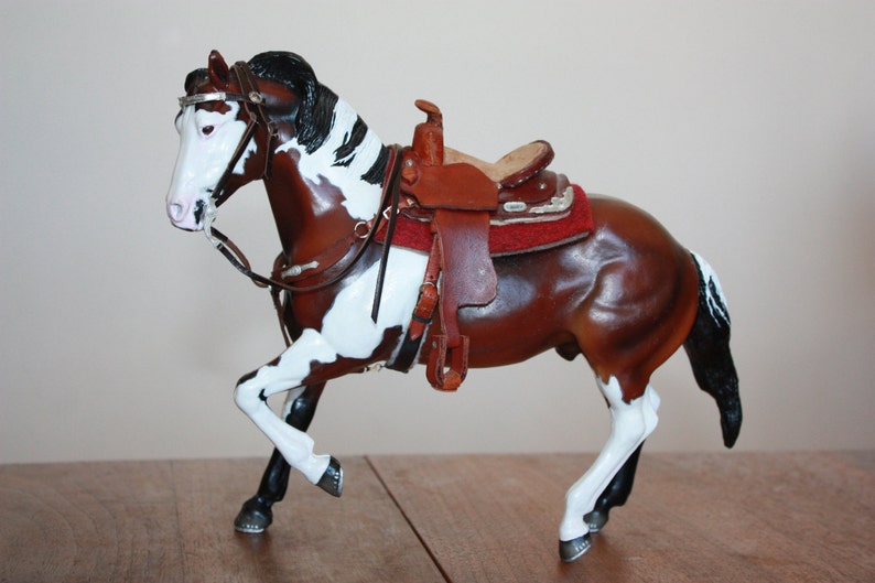 Puede incluir: Una figurita de caballo pintada de marr&oacute;n y blanco con una crin y cola negras. El caballo lleva una silla de montar de cuero marr&oacute;n con una almohadilla roja y detalles plateados.