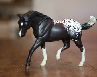 Puede incluir: Una figurita de caballo manchado de blanco y negro con una crin y cola negras. El caballo está de pie sobre sus cuatro patas y tiene una mancha blanca en la cara.