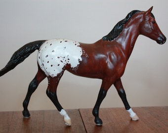 Puede incluir: Una figurita de caballo marrón y blanco con manchas, con una crin y cola negras. El caballo está de pie sobre sus cuatro patas y está en una pose de caminar.