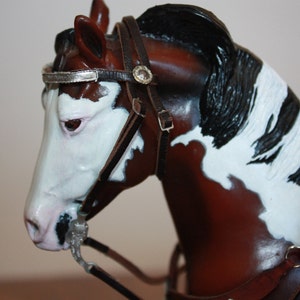 Puede incluir: Primer plano de una figurita de caballo pintada de marr&oacute;n y blanco que lleva una brida de cuero negro con detalles plateados. El caballo tiene una crin y una cola negras.