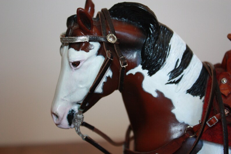 Puede incluir: Primer plano de una figurita de caballo pintada de marr&oacute;n y blanco que lleva una brida de cuero negro con detalles plateados. El caballo tiene una crin y una cola negras.