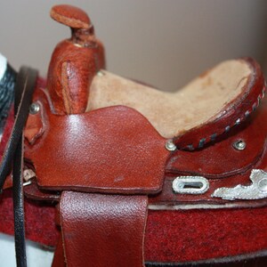 Puede incluir: Una silla de montar de cuero marr&oacute;n con detalles plateados y un asiento de fieltro rojo. La silla est&aacute; unida a un caballo blanco y negro.